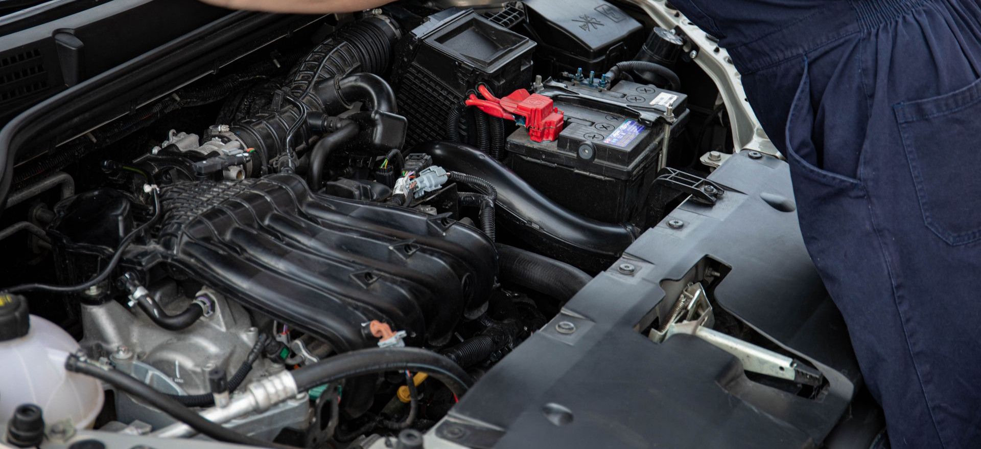 Mechanic performing maintenance on a car engine outdoors during the day.